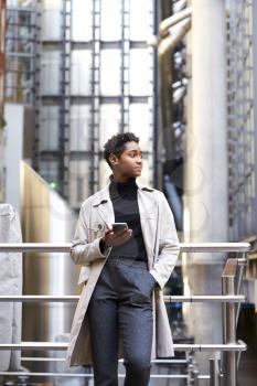 Fashionable young black woman standing in the city leaning on a handrail holding smartphone, low angle, vertical