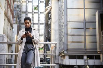 Fashionable young black woman standing in the city leaning on a hand rail using her smartphone, low angle