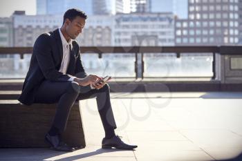 Millennial black businessman wearing black suit and white shirt sitting on the Thames embankment, London, using smartphone, backlit