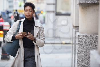 Young black businesswoman walking in the street in London using her smartphone, front view