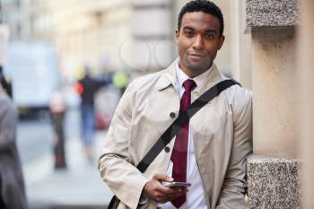 Young black businessman leaning on a wall in the street in London holding phone and smiling to camera, selective focus