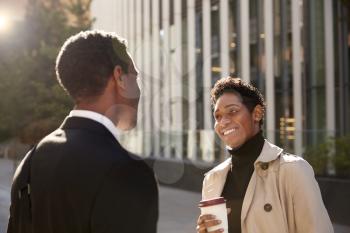Smiling young black businesswoman standing on the street with a takeaway coffee, talking to her male colleague, selective focus