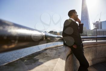 Young black businessman wearing a suit leaning on a wall at Thames embankment, London, using smartphone, low angle