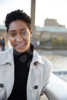 Young black woman wearing a turtleneck sweater and a mackintosh standing on Millennium Bridge, London, looking to camera smiling, vertical