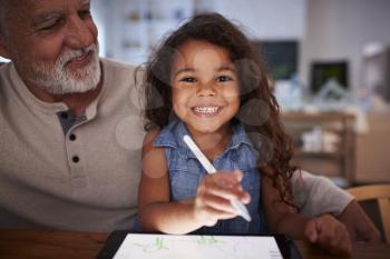 Senior Hispanic man with his young granddaughter using stylus and tablet computer, smiling to camera