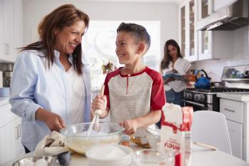 Pre-teen boy making a cake in the kitchen with his grandmother, his mum standing in the background