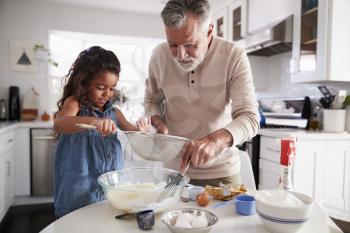 Young girl preparing cake mixture with her grandfather at the kitchen table, close up