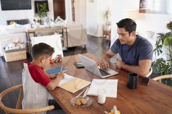Hispanic father and son working opposite each other at the dining room table, elevated view
