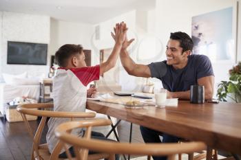 Hispanic father and son sitting opposite each other, high five over the dining room table, side view