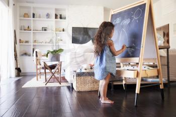 Young Hispanic girl drawing with chalks at a blackboard in the living room at home, full length
