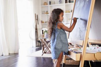 Young Hispanic girl drawing with chalks at a blackboard in the living room at home, side view