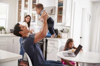 Young Hispanic family in their kitchen, dad lifting baby in the air, mum cooking at hob, close up