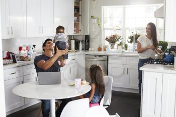 Young Hispanic family in their kitchen, dad holding baby in the air, mum cooking at the hob