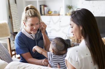 Female healthcare worker visiting young mum and her infant son at home, using stethoscope, close up