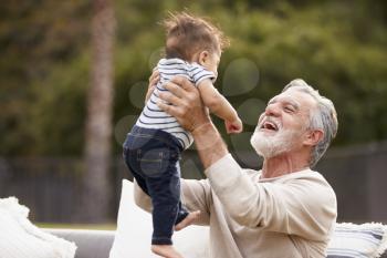 Senior Hispanic man sitting in the garden lifting his baby grandson in the air and smiling to him