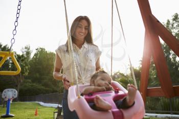 Young Hispanic mother pushing her baby on a swing at a playground in the park, close up