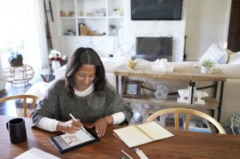 Elevated view of middle aged mixed race woman sitting at a table in her dining room using a stylus with a tablet computer, elevated view, focus on foreground