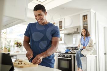 Young adult man standing in the kitchen preparing food, his partner sitting on kitchen worktop behind him, focus on foreground
