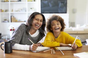 Middle aged mixed race woman sitting at table in her dining room drawing with her pre-teen granddaughter, laughing to camera, front view, close up