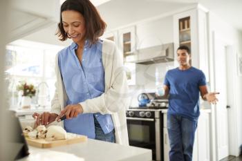 Young mixed race woman standing in the kitchen preparing food, her partner standing behind talking, focus on foreground