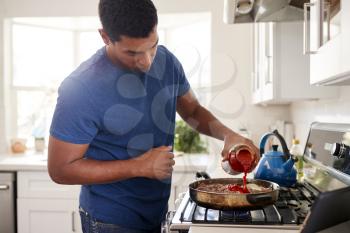 Young adult black man standing in the kitchen cooking on the hob, adding a sauce to the frying pan, close up