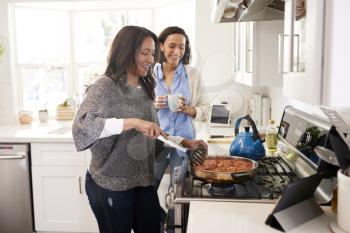 Middle aged woman standing in the kitchen cooking at hob following recipe on a tablet computer, her adult daughter standing beside her talking, side view