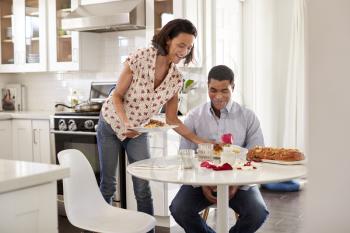 Young adult man sitting at the table in the kitchen is served a romantic meal by his partner, selective focus