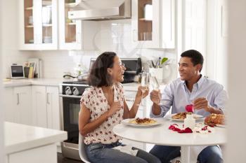 Young couple sitting at the table in their kitchen making a toast while eating a romantic meal together, selective focus