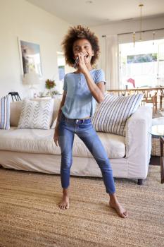 Pre-teen black girl dancing and singing in the living room at home using her phone as a microphone, full length, vertical