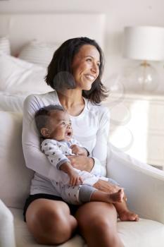 Young adult mother sitting in an armchair in her bedroom, holding her three month old baby son in her arms and looking away smiling, vertical