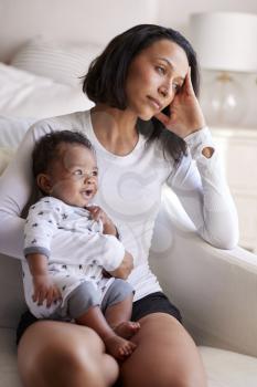 Young adult mother sitting in an armchair in her bedroom, holding her three month old baby son, looking away in contemplation, vertical