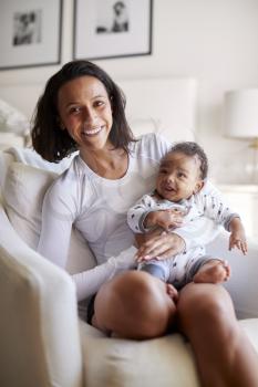 Happy mixed race young adult mother sitting on an armchair holding her three month old baby son, smiling to camera, vertical