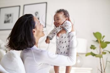 Happy mixed race young adult mother raising her three month old baby son in the air, smiling at him, close up