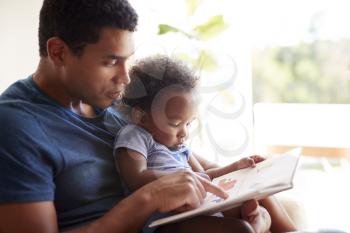 Close up of young adult black father reading a book with his two year old son, close up, side view, backlit