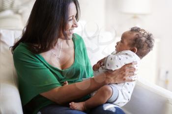 Middle aged mixed race grandmother sitting in an armchair holding her three month old grandson and smiling, focus on foreground