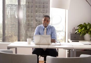 Portrait Of Male Financial Advisor In Modern Office Sitting At Desk Working On Laptop