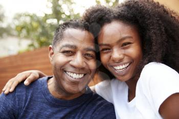 Middle aged black dad and teenage daughter embracing and smiling to camera, close up