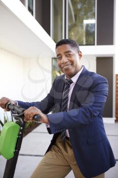 Smartly dressed middle aged black man standing on electric scooter in front of his house, close up