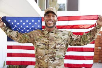 Millennial black soldier standing outside modern building holding US flag, smiling to camera, close up