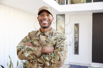 Soldier in camouflage standing outside modern house with arms crossed smiling to camera, close up