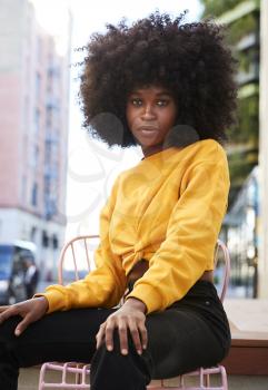 Young black woman with afro hair and sitting on a chair in the street looking to camera, vertical