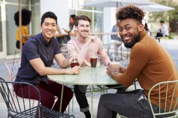 Three millennial male friends talking over cold brews outside a cafe in a city street turn to camera