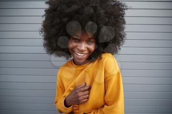 Young black woman with afro against grey wall, raising shoulder, smiling to camera, close up