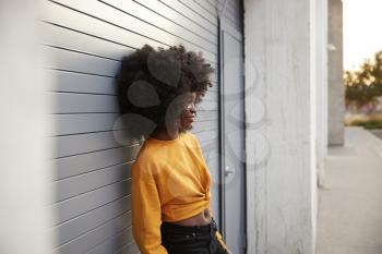 Young black woman with afro leaning in the street against grey security shutters, side view