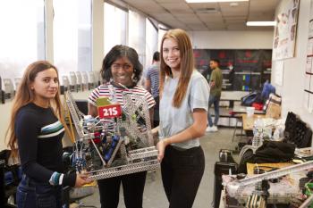 Portrait Of Female University Students Carrying Machine In Science Robotics Or Engineering Class