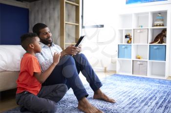 Father With Son Sitting In Bedroom Playing Game On Digital Tablet Together