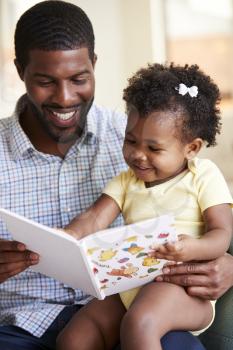 Baby Daughter And Father Sitting On Sofa Reading Book Together