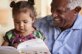 Grandfather Sitting On Sofa At Home With Baby Granddaughter Reading Book Together