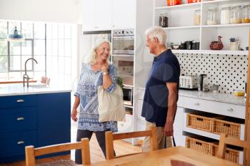 Senior Couple Returning Home From Shopping Trip Carrying Grocery Bags Through Kitchen
