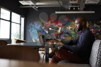 Mid adult black male creative sitting at table in office dining area using a laptop, side view
